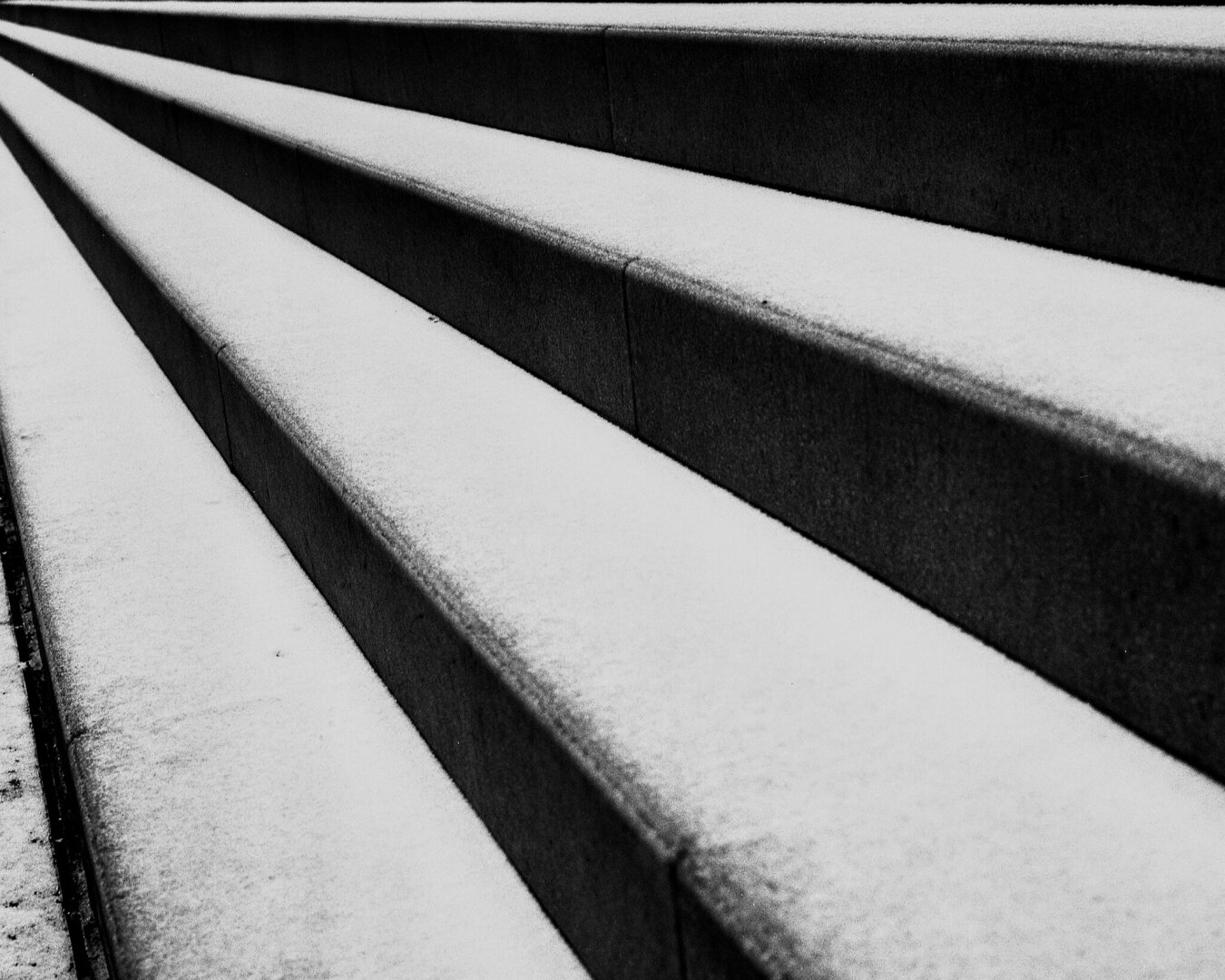 The image shows a series of stone steps covered in a layer of snow. The steps are arranged in a diagonal pattern, with the foreground featuring the top edge of the steps and the background fading into more steps. The image is in black and white, emphasizing the texture of the snow and stone.