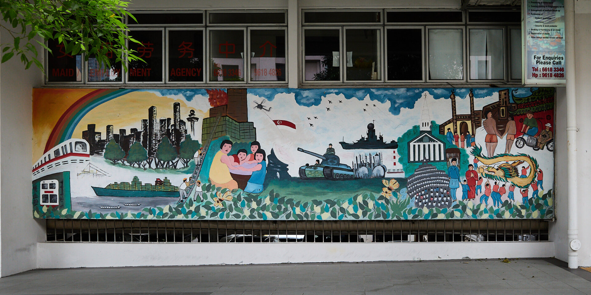 A large mural covers a wall, featuring various scenes including a train, a cargo ship, and a tank. People are depicted in different activities, with a rainbow arching above. The background includes buildings, trees, and a flag. There are also windows above the mural with red text, possibly indicating a business.
