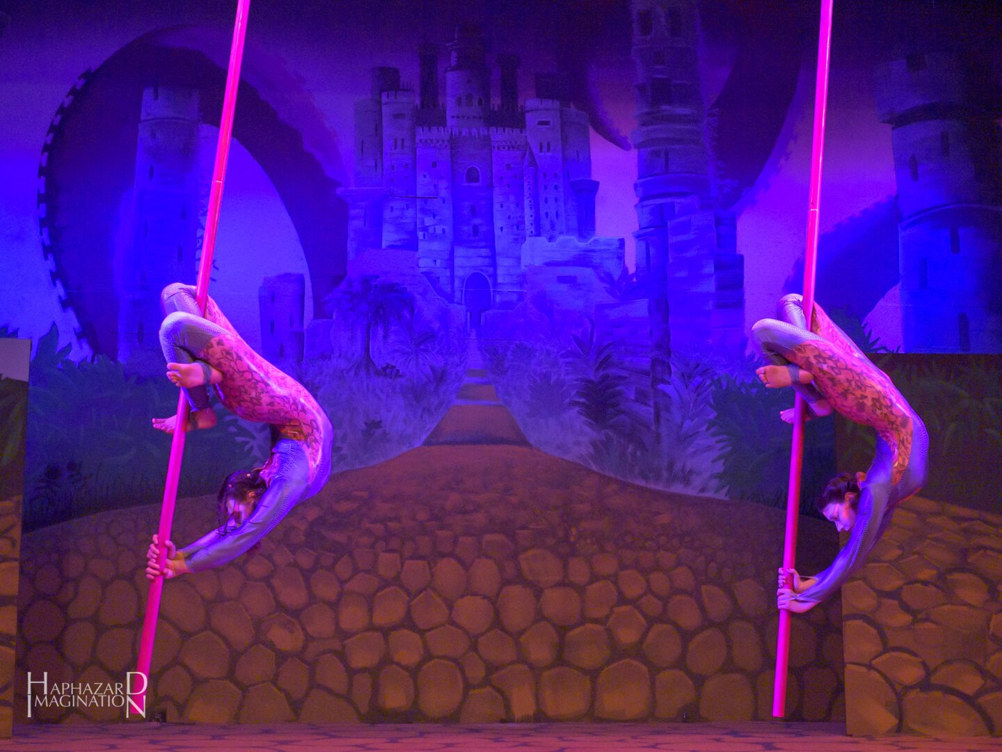 Two aerial pole artists perform back bends on flying pole, wearing colorful outfits against a castle and courtyard background.