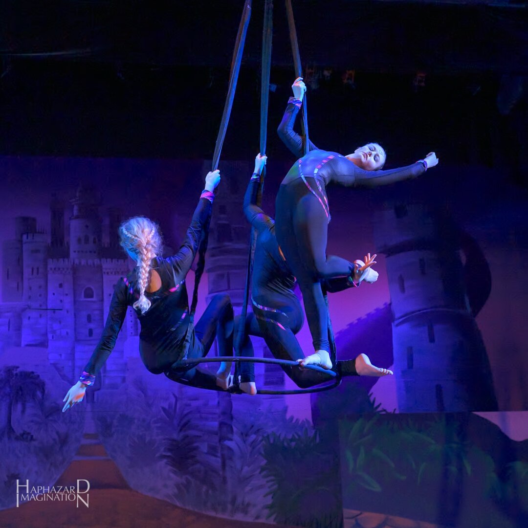 Three aerialists are on a specially rigged lyra making artistic poses. They are wearing mostly black costumes.