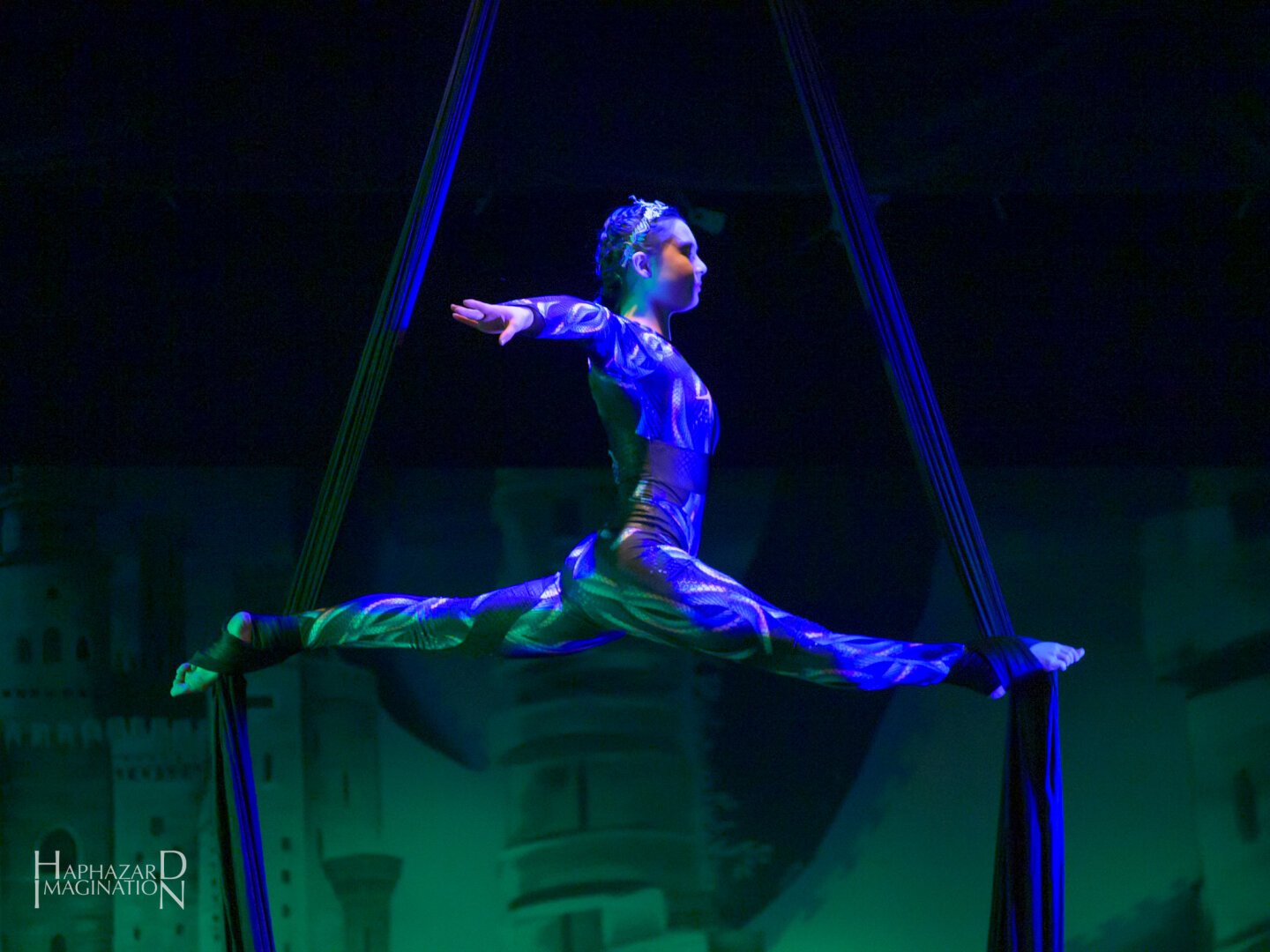 An aerial fabric artist in a perfect split balance, shown in profile. She is wearing a blue and white patterned costume, with dark blue silks and there green list castle on the backdrop behind her.