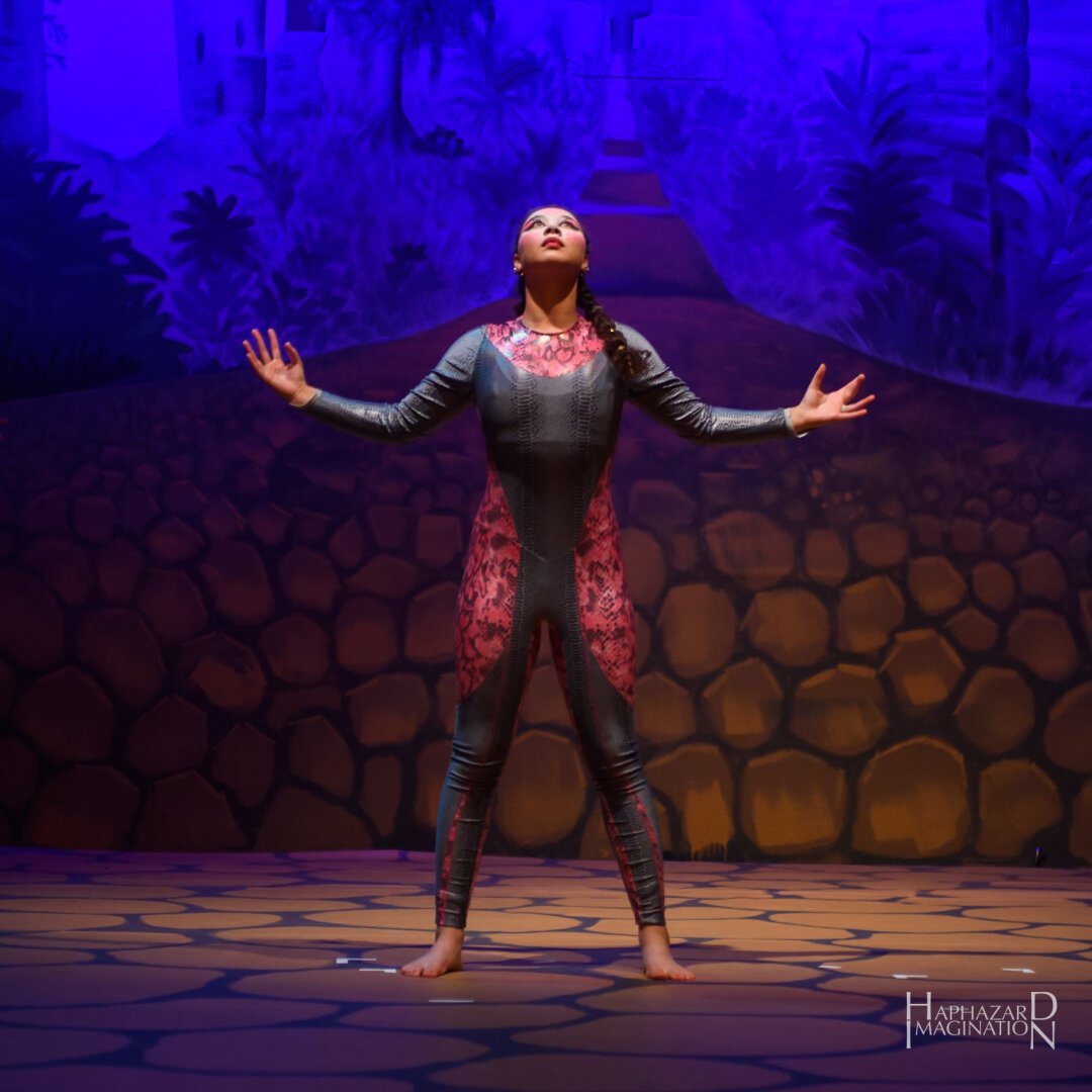 Circus performer in grey and red tights raises their arms and looks towards the sky. In the background is a path leading to a castle.