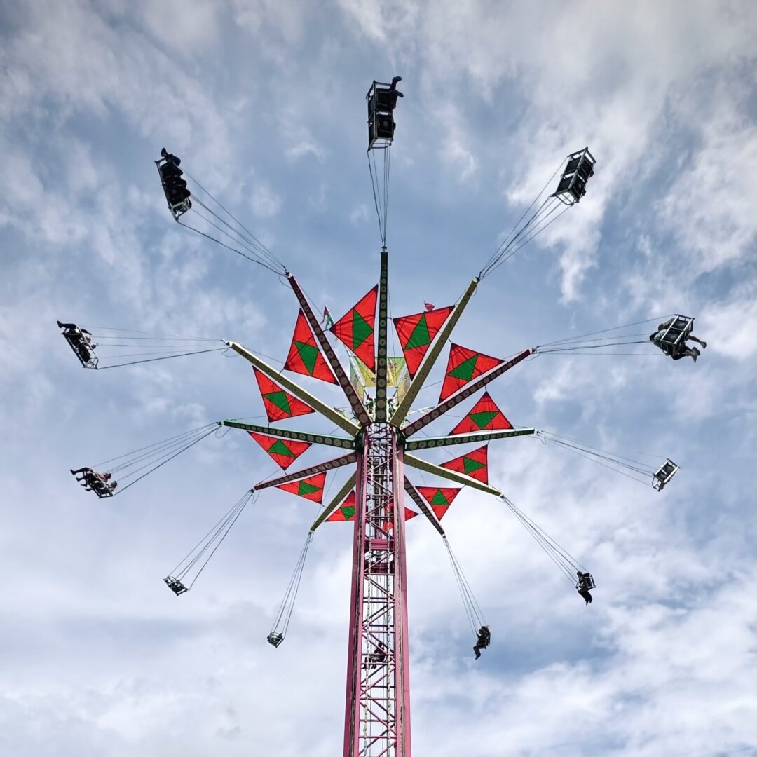 A fair's half-full giant swing ride is seen from below on a mostly sunny day.
