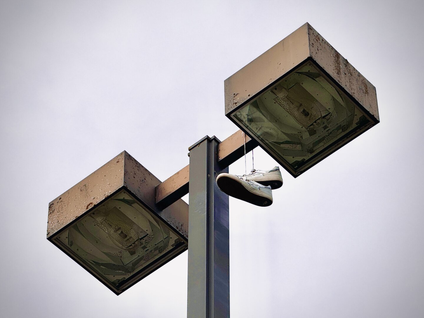 White skate shoes hang by their laces from a rusty metal street lamp under a cloudy sky.