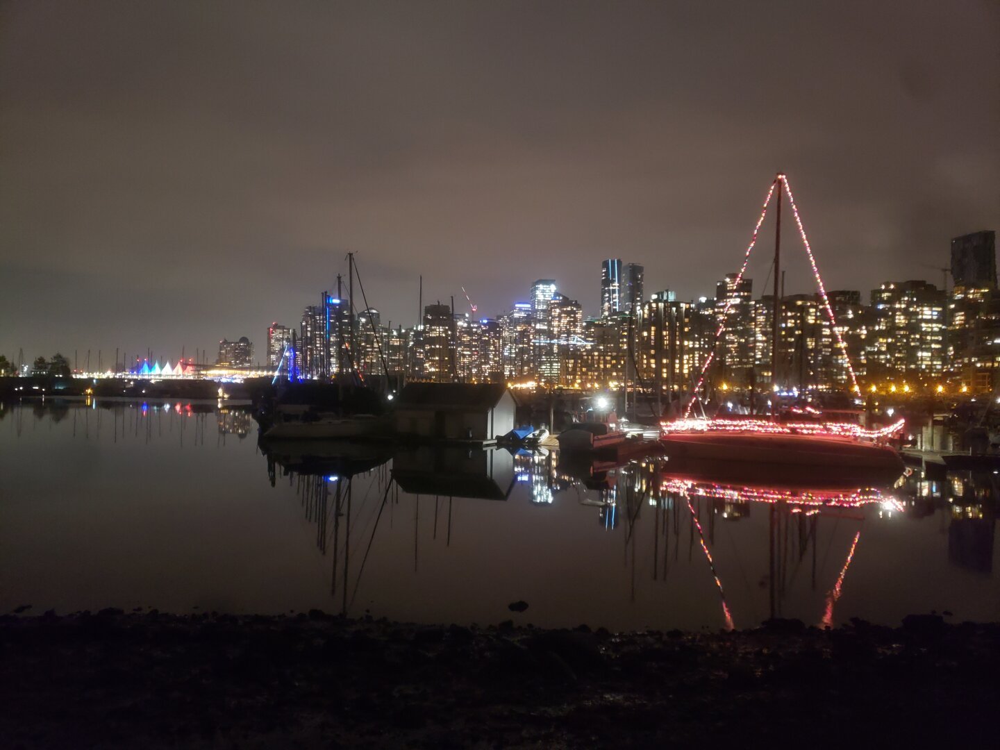 A night-time picture of the North West side of the downtown Vancouver skyline. There are decorated boats in the marina in the foreground, and light spills out from the streets and buildings in the background.