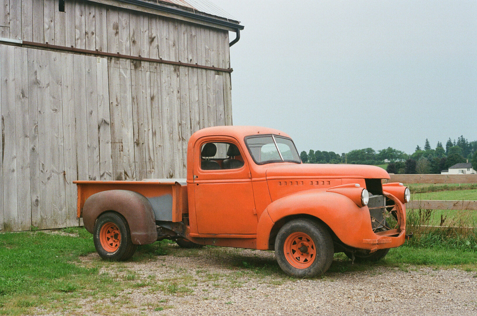 An orange '46 chevy truck with some mismatched parts, parked by an old weathered barn. There are some rolling hills and trees in the distance, and a grey overcast sky.