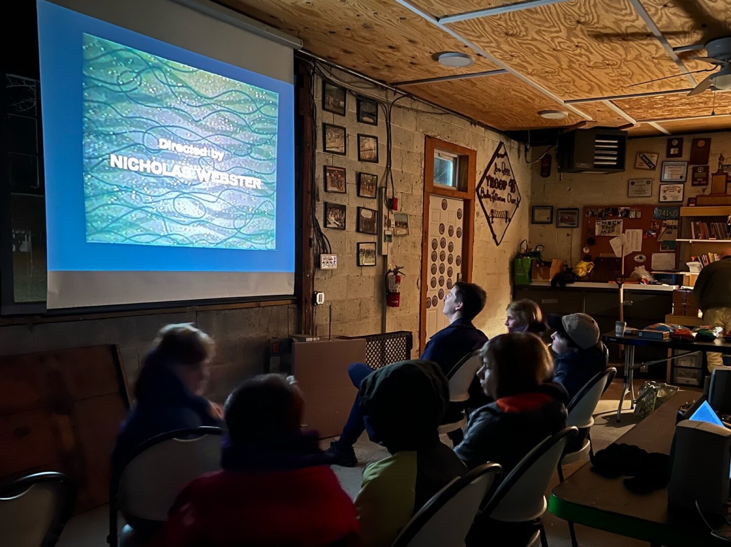 Photo of kids in chairs watching a movie on a screen inside a shed.