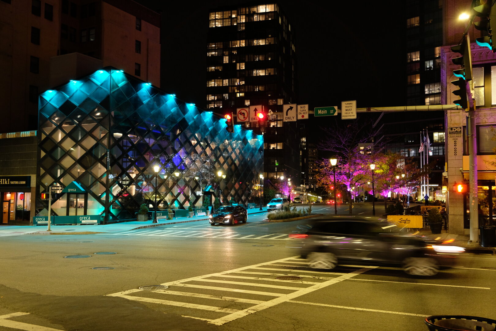 A downtown White Plains intersection at night. A car has passed through the intersection, moving from left to right. It is noticeably blurred and clearly in motion. There are colorful blue lights on the facade of a building at the intersection. Behind it is a much taller building, likely a condominium. 
On the other side of the street are numerous trees that have lost many of their leaves. The ones that remain are purple (lit from below by purple lights).
A "Main ST" sign hangs above the intersection along with a traffic light and some street signs.