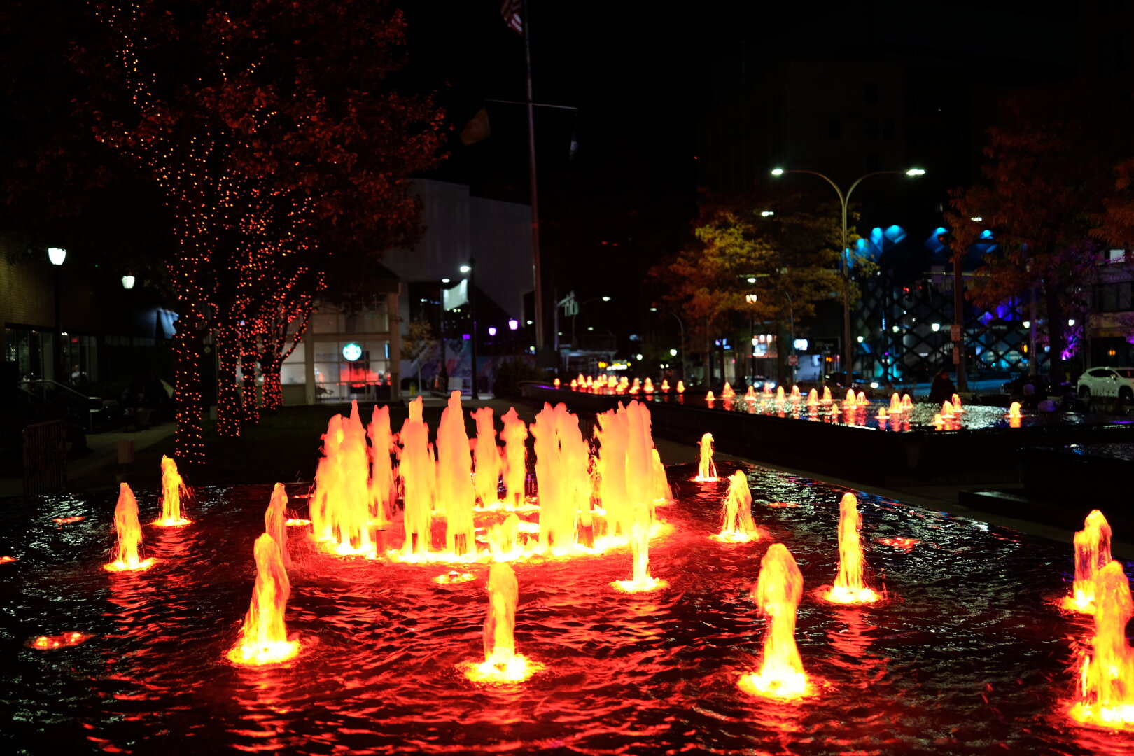 An elevated fountain at night. It is about waist-high. The water spouts are arranged in a somewhat concentric circle pattern, gently spouting water about a foot above the water line. Each one is lit from below in a red glow.
Trees adorned with red lights lie behind the fountain to the left.