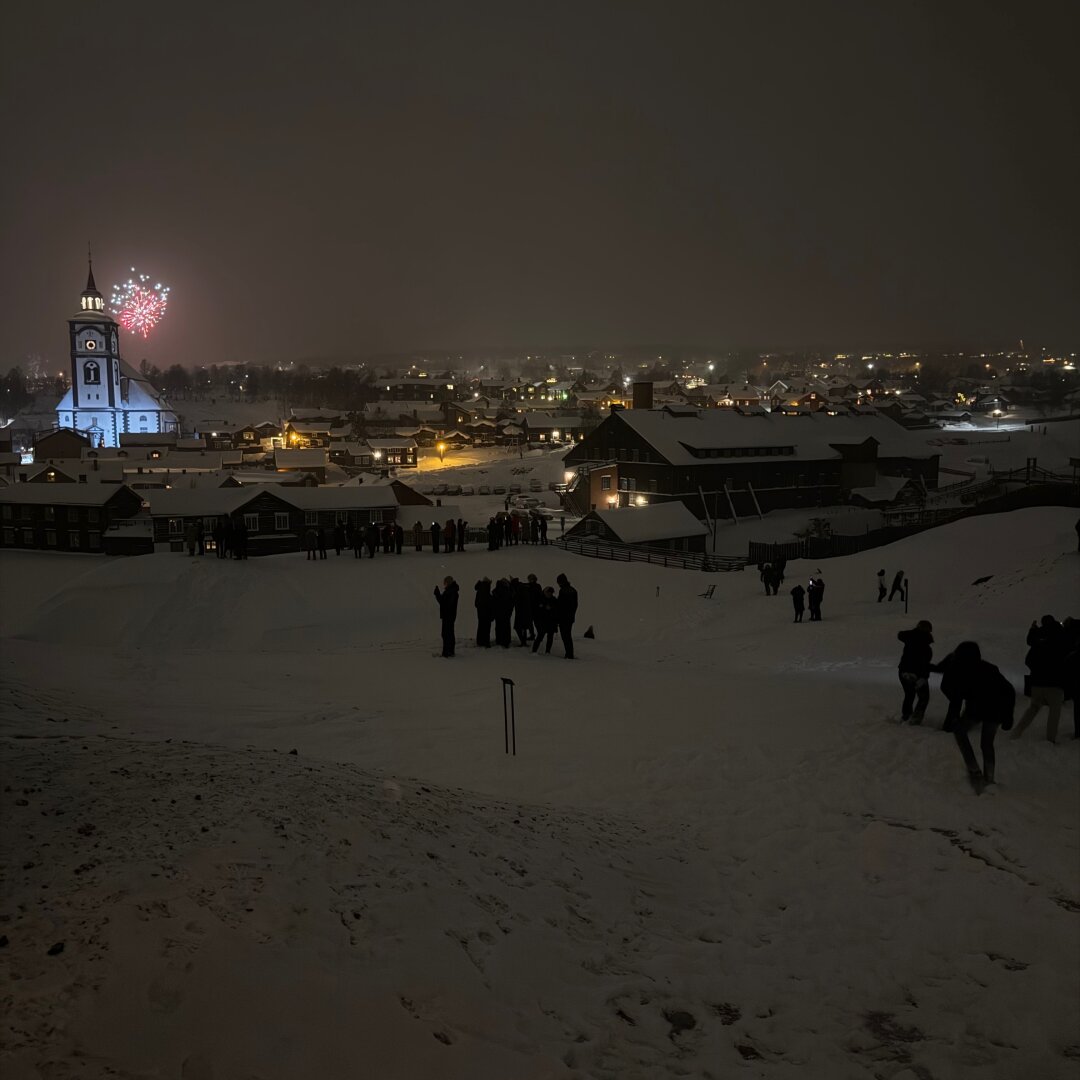 Opplyst Røros kirke til venstre med fyrverkeri bak kirketårnet. Det er eller mørkt men man kan skimte folk som står i snøen.