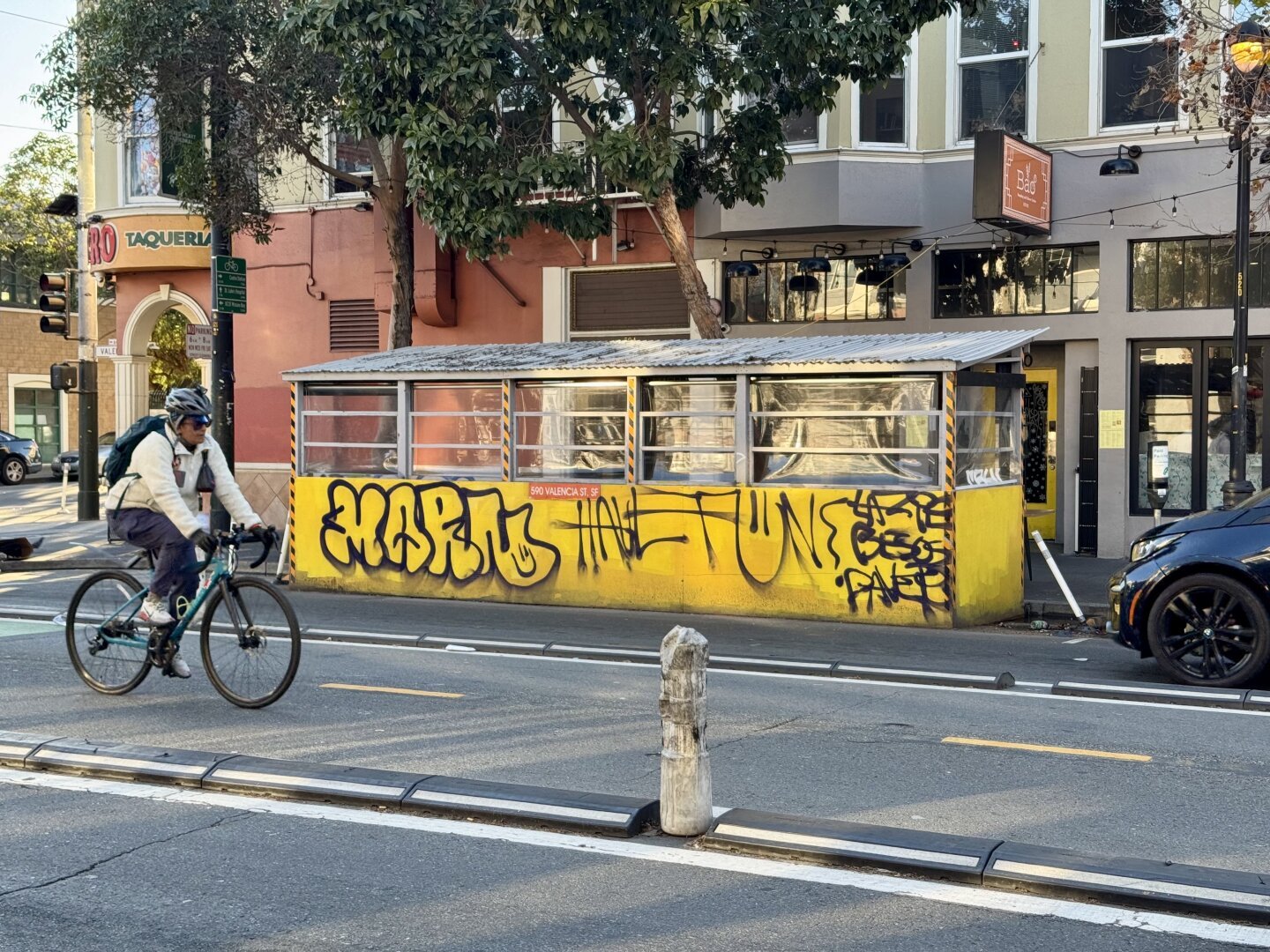 A shot looking across Valencia Street in San Francisco with a bicyclist riding in the foreground. The main focus is on a yellow parklet with graffiti on it that says “Marn” and “Have Fun” in black spray paint.