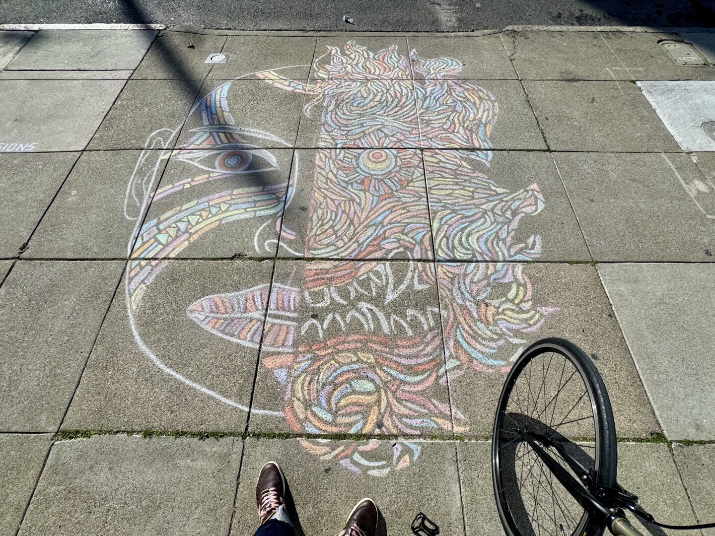 A large piece of art drawn on the sidewalk in chalk. The piece is by Chalk Visions and is face. It's about 4 feet wide and 4 feet tall. Half of the face is normal, the other half looks deformed, maybe like a demon. It sort of looks like Two-Face, the character from Batman, but I don't think that's actually who is being drawn here. The colors are pink, yellow, and light blue.