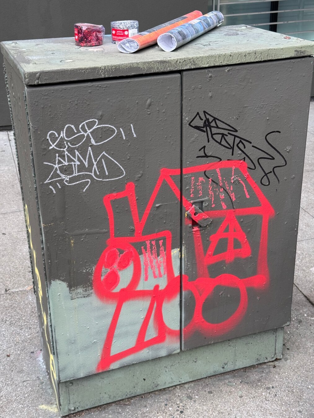 A utility box with some graffiti tags on it. One big one is a drawing of a train. It was painted with red spray paint, and there is no signature. The train sort of looks like Thomas the Tank Engine or some kid's book drawing of a train.