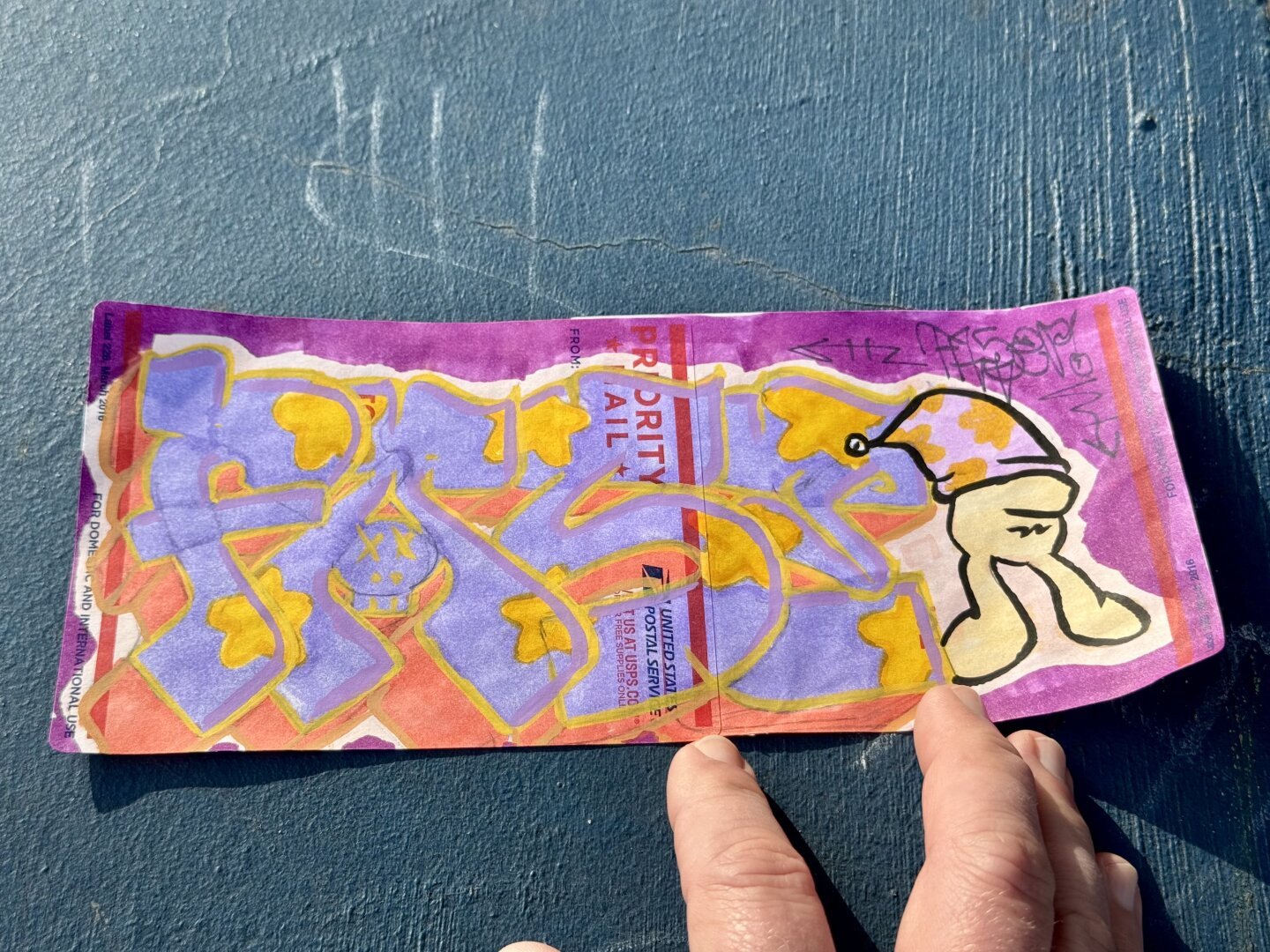 Two usps priority mail stickers that have been attached to each other to make a long sticker. It has art on it that says something like “Fast” I. A graffiti style with purple fill and yellow stars. There is also a drawing of something that looks like Cheech Wizard. The sticker has lost its stickiness, so it’s laying on a surface, but not stuck to it.