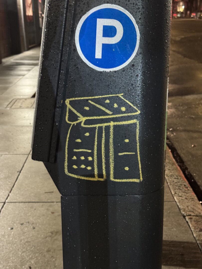 Graffiti on a parking meter. The graf is a drawing of three dominoes in yellow paint marker. Two of the dominoes are stacked in vertical columns with the third one laying across the top.