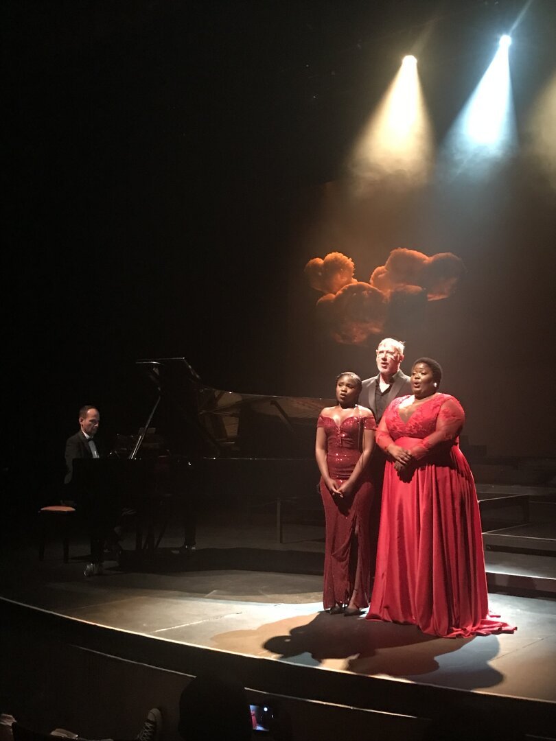 Picture  of a stage, lit in red and black hues. Taken of three singers, Professor Frederico Freschi (back) and Khanyakazi Madlala (right) and Sisikelewe Mngenela (left) performing. Grand Piano included.