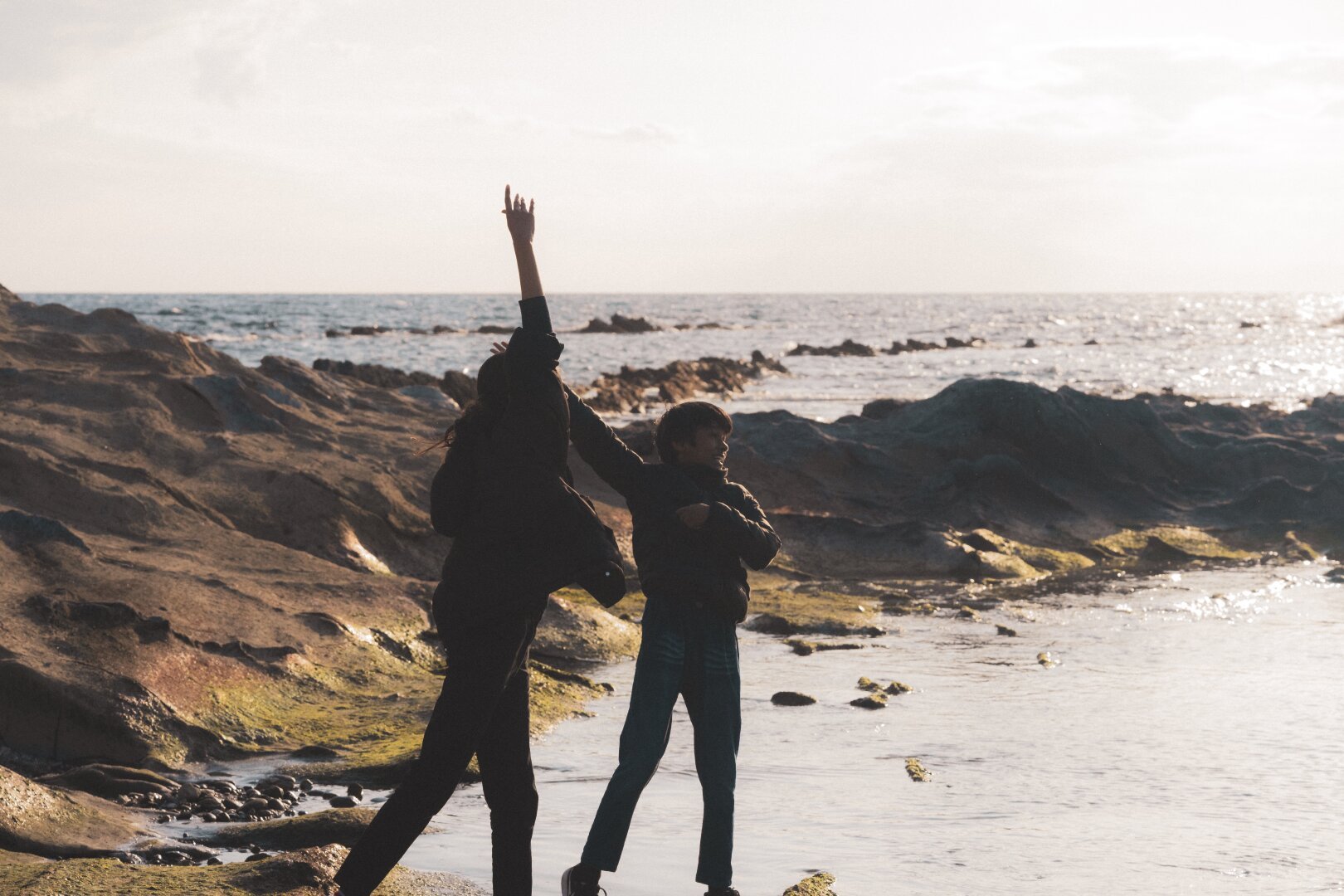 The image captures two silhouetted figures against a coastal backdrop during sunset. The adult is raising their hand towards the sky as if reaching out to it, while the child mimics the gesture with both hands up. They stand on rocky terrain near the water's edge where small pools of shallow waters reflect light from the sun-drenched surface. Beyond them stretches a vast expanse of sea meeting the horizon under a soft gradient of colors in the sky, suggesting an overcast day. The muted tones and elongated shadows hint at a low-angle sunlight typical of dawn or dusk hours. No distinct facial features are discernible due to backlighting, preserving anonymity for the subjects depicted.