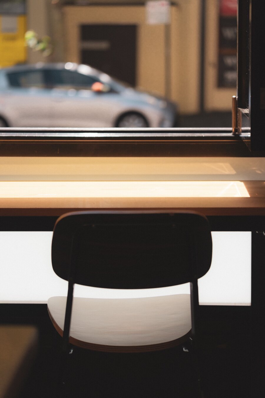 The image captures an interior scene with a focus on a solitary black chair positioned at a table near a large window that allows natural light to fill the space. Through the glass panes, we can observe a parked car outside, suggesting an urban environment or possibly a parking lot adjacent to this establishment. The blurred background hints at more cars and perhaps buildings, giving context but not being in focus enough for detailed analysis. Inside, on the table is what appears to be a white lampshade with no visible light source beneath it, indicating that either the bulb is off or behind view of our perspective. The colors are muted with earthy tones dominating the scene, highlighted by the gleaming surface of the polished wood and glass.