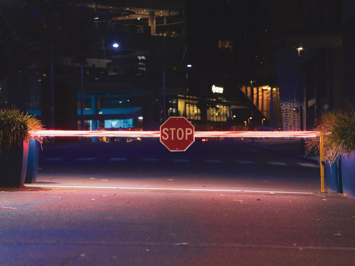 The image captures an urban nighttime scene where red light streaks across the frame, suggesting movement from vehicles. In the foreground is a prominent 'STOP' sign mounted on two posts, positioned to block traffic flow. The background features what appears to be city infrastructure such as buildings and possibly elevated roads or parking structures with glowing lights. Trees are visible in small planters on either side of the stop barrier, adding some greenery to this urban setting.