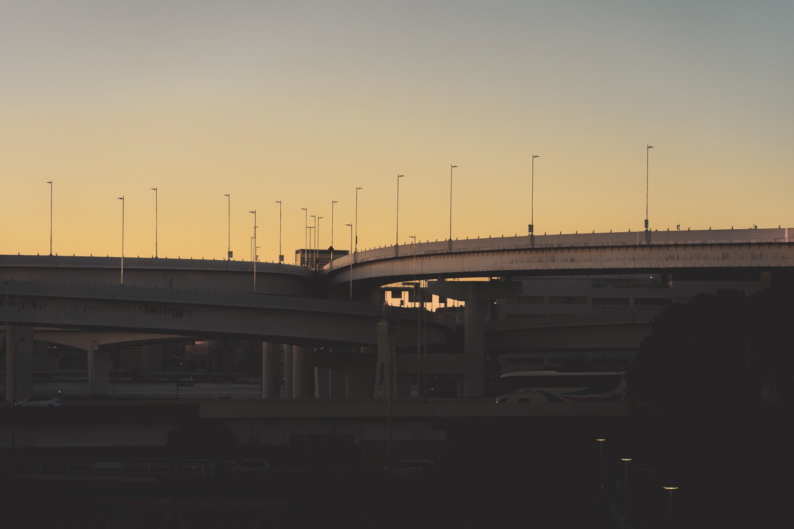 The image depicts an urban landscape during twilight hours with artificial lighting beginning to illuminate the scene. The main subject is a complex network of elevated roadways and bridges that weave over each other, indicative of a bustling metropolitan area. Silhouettes suggest traffic flow on these roads as they cut across the sky at various angles. Street lamps stand sentinel along the edges, their lights just starting to glow against the fading natural light in the dusky sky above.

The color palette is dominated by muted tones, with the roadways and structures rendered in dark shades contrasted sharply against the warm hues of sunset—a gradient ranging from pale yellow near the horizon to deeper orange higher up. The background's twilight lends a serene yet busy ambiance to an otherwise industrial scene. No people or moving vehicles are discernible due to the silhouette effect caused by backlighting, suggesting either early morning or late evening timing.