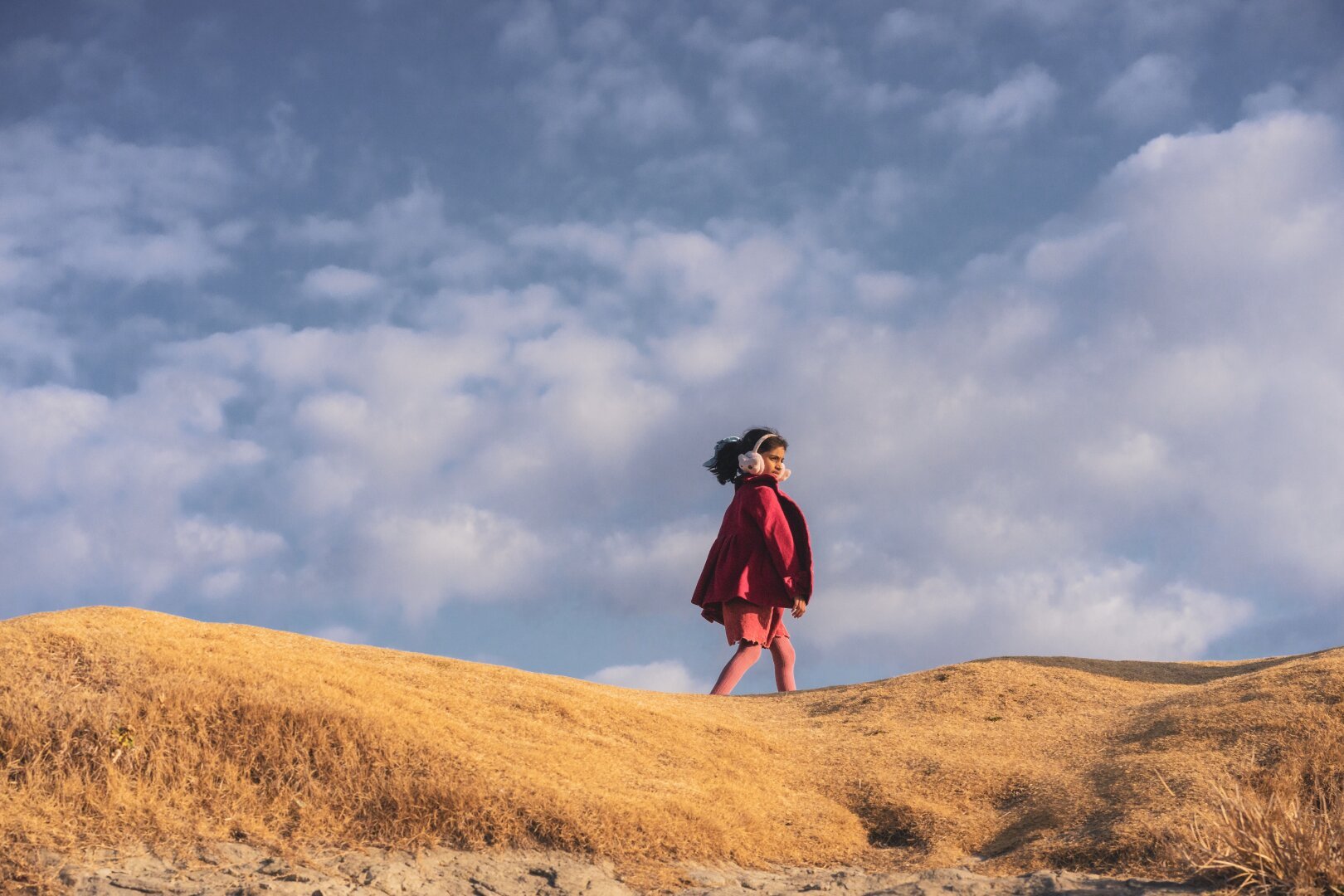 The image depicts an individual standing atop what appears to be a dry grass-covered hill under a partly cloudy sky. The person is dressed in vibrant red attire that includes a coat and pants, with pink leg warmers or leggings visible beneath the pants. They are wearing earmuffs on their head and have long hair trailing down behind them as they look ahead towards the horizon. Their posture suggests movement forward along the slope of the hillside. In terms of color contrast, the reds stand out against the golden tones of the grassy terrain and the cool blues of the sky above. The setting conveys a sense of solitude or introspection amidst nature's expansive backdrop.