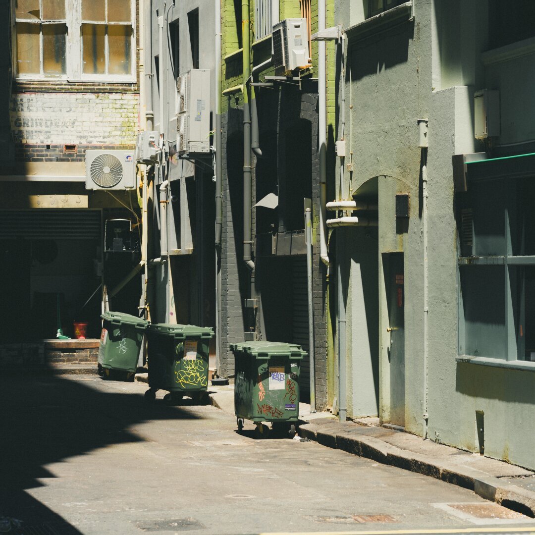 The image captures an urban alleyway during daylight with bright sunlight casting shadows on the ground. In the foreground are several green garbage bins lined up against the buildings, some displaying graffiti and stickers. The middle ground reveals more architectural details such as weathered brick walls with visible deterioration, air conditioning units attached to the exterior of a building, and various pipes running along the sides. A vent is also seen on one of the walls. In the background, windows reflecting sunlight suggest an adjacent structure not fully captured in the frame. The overall color palette consists of muted urban tones—greys of the pavement, green from the bins, and faded brick reds and yellows from the buildings' façades. There are no people visible in this scene, suggesting a quiet or deserted moment within what appears to be an industrial or less-frequented part of town.