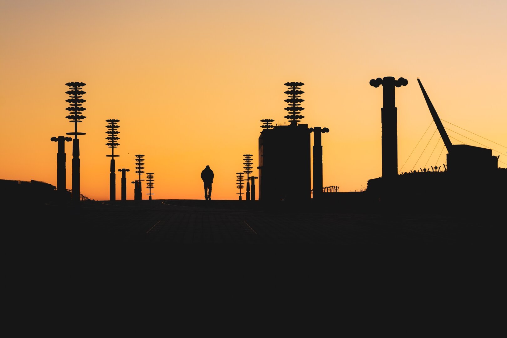 The image depicts an urban scene at twilight with silhouetted figures against a gradient sky transitioning from orange to yellow near the horizon and darker shades as it extends upward. To the right stands an industrial structure that resembles part of a bridge's support system, characterized by its towering pillars and overhead cables converging towards one end where they are likely attached to another segment out of view. The overall atmosphere is serene yet conveys the bustle of city infrastructure as it recedes into nightfall.