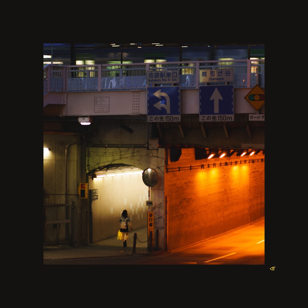 Night street scene under an overpass in Japan, with Japanese road signs above a tunnel entrance; a lone pedestrian stands near a closed shuttered doorway on the left, while the tunnel on the right is lit orange by overhead lights.