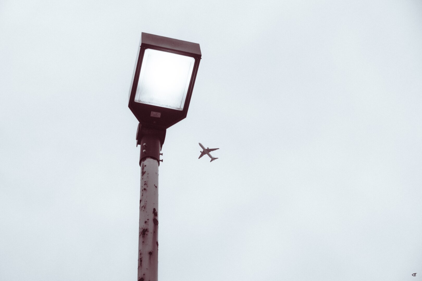 Tall streetlight with a glowing square lamp on the left, viewed from below against a pale overcast sky, with a small airplane flying in the distance near the center.