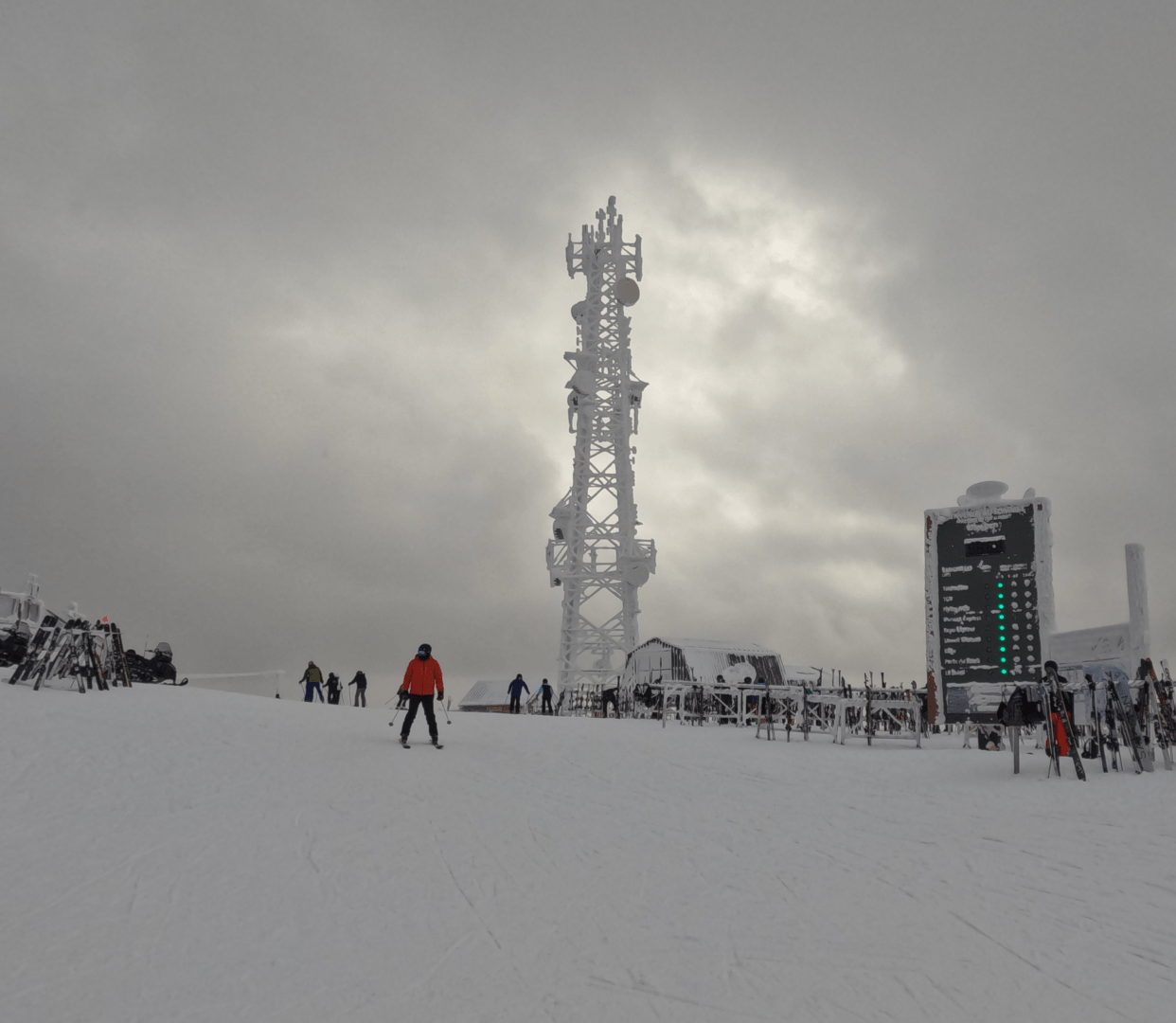 snowy landscape, the whole image is white because of the white snow and the white gray sky. there is a frozen communication tower in the middle of the picture and skiers all around