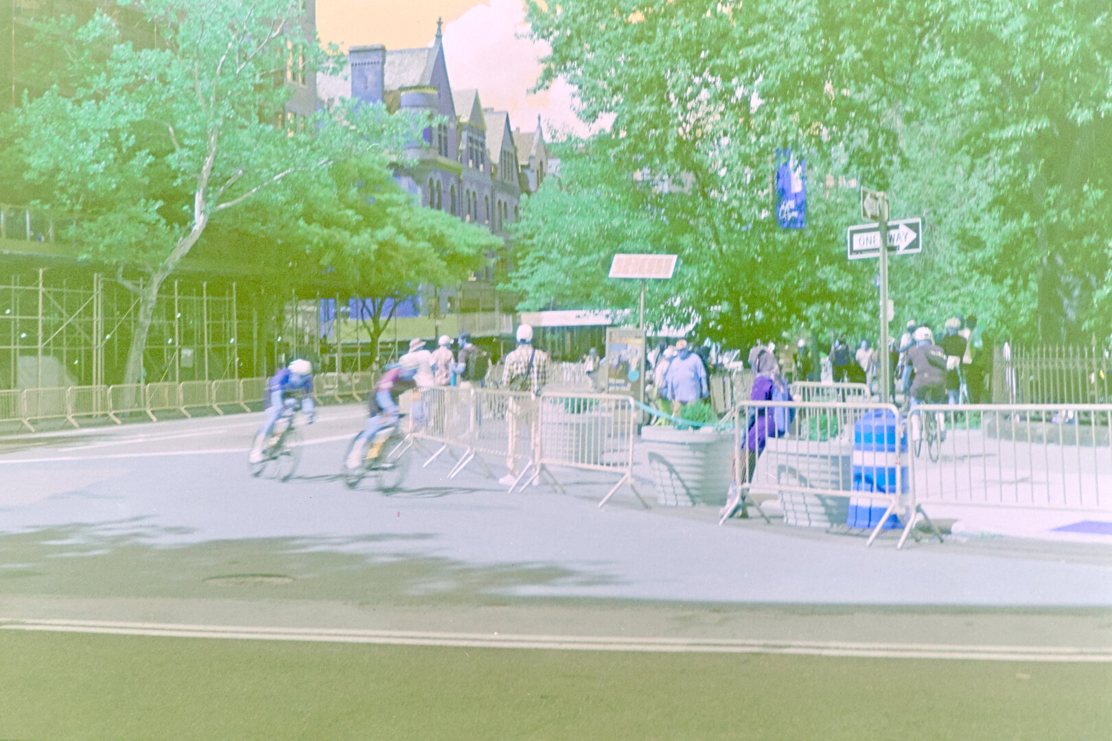 Two cyclists making a turn at high speed, leaning in, with motion blur. In the background are many trees and old houses. The funky film turned the sky orange and skin tone blue.