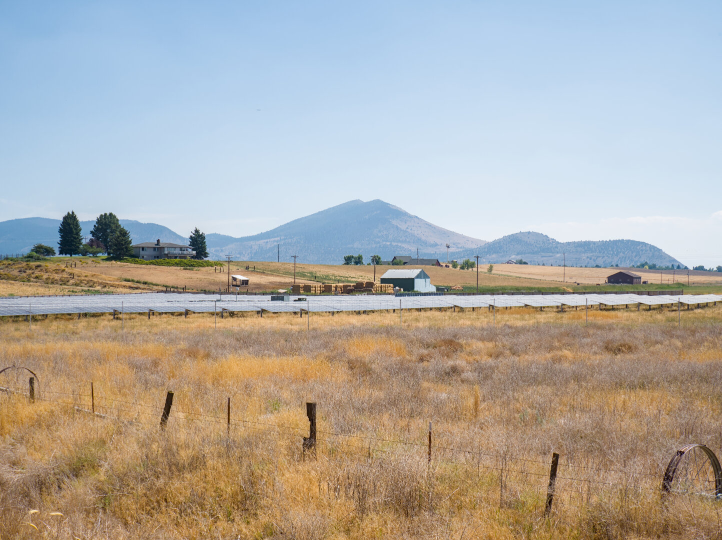 A dry yellow field with solar panels, with a little hill with a house and trees on it and mountains in the distance
