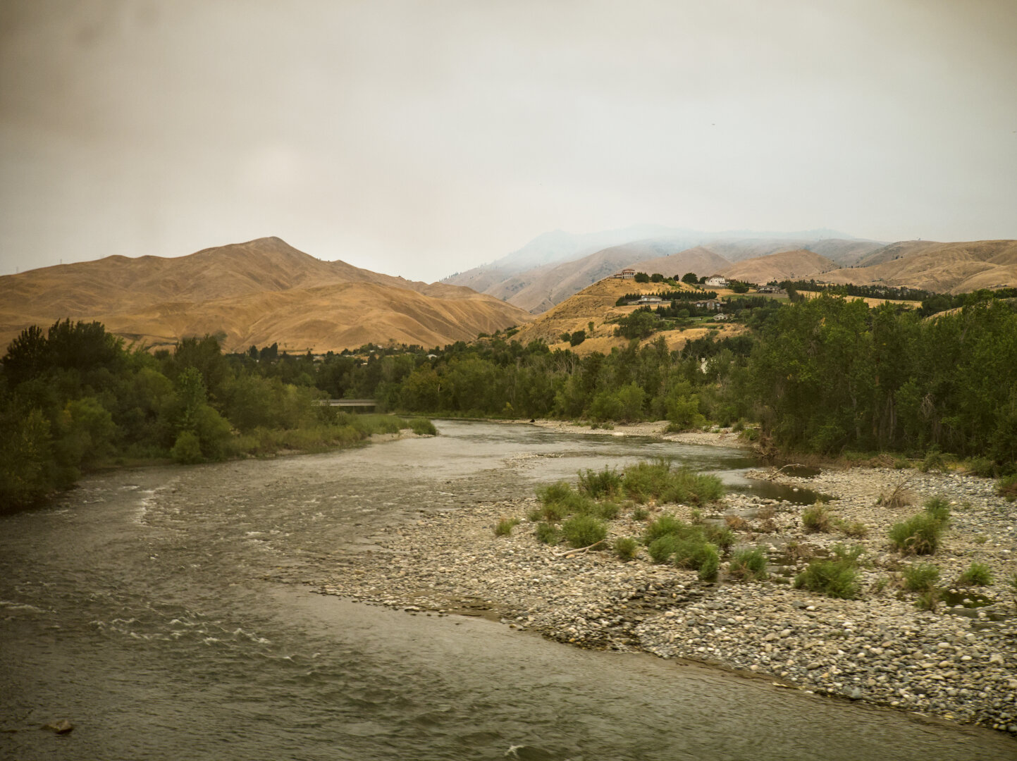 A large and shallow river winds into the scene towards many dry hills covered by yellow grass. There's a forest surrounding the river. The sky was hazy and misty, orange from the wildfire.