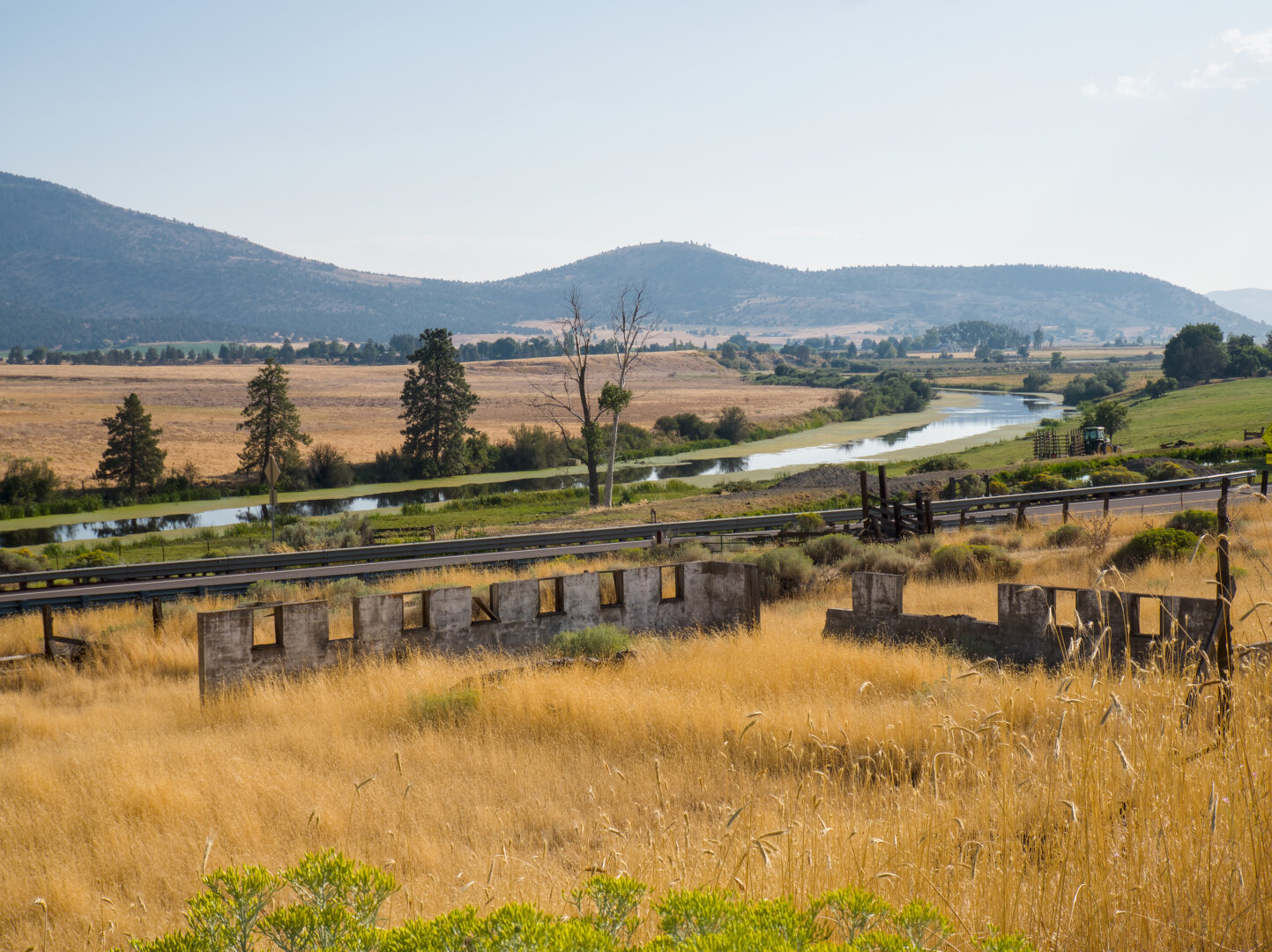 Looking down a hill covered in dry yellow grass as the ruins of a small concrete building. Beneath the hill is road and a river. Across the river is a dry yellow field and some mountains.
