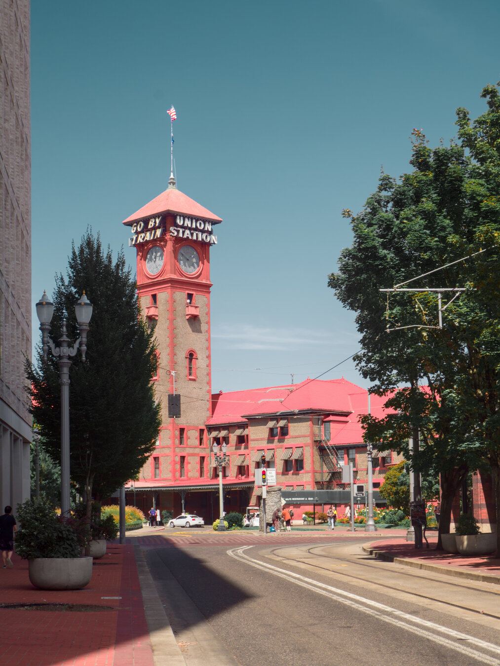 A small street with trolley tracks lead to a Romanesque-inspired red brick train station building, framed by trees on both sides. Above the clock of the clock tower are bold white Italic letter saying "Go by train" on one side and "Union Station" on the other side.