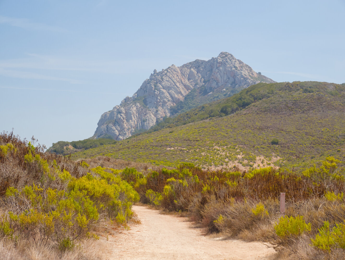 A rugged rocky peak rises above gentler slopes covered in shrubs. There's a dirt trail in the foreground.