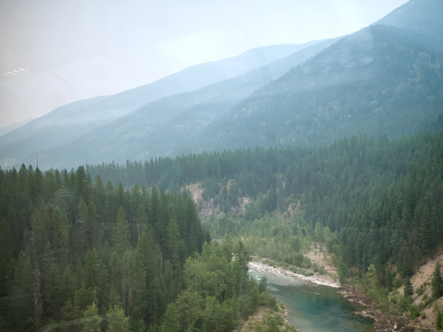 A large river flows beneath mountains that are covered in conifer forests. It was cloudy and misty. Reflection of the observation car windows can be faintly seen.