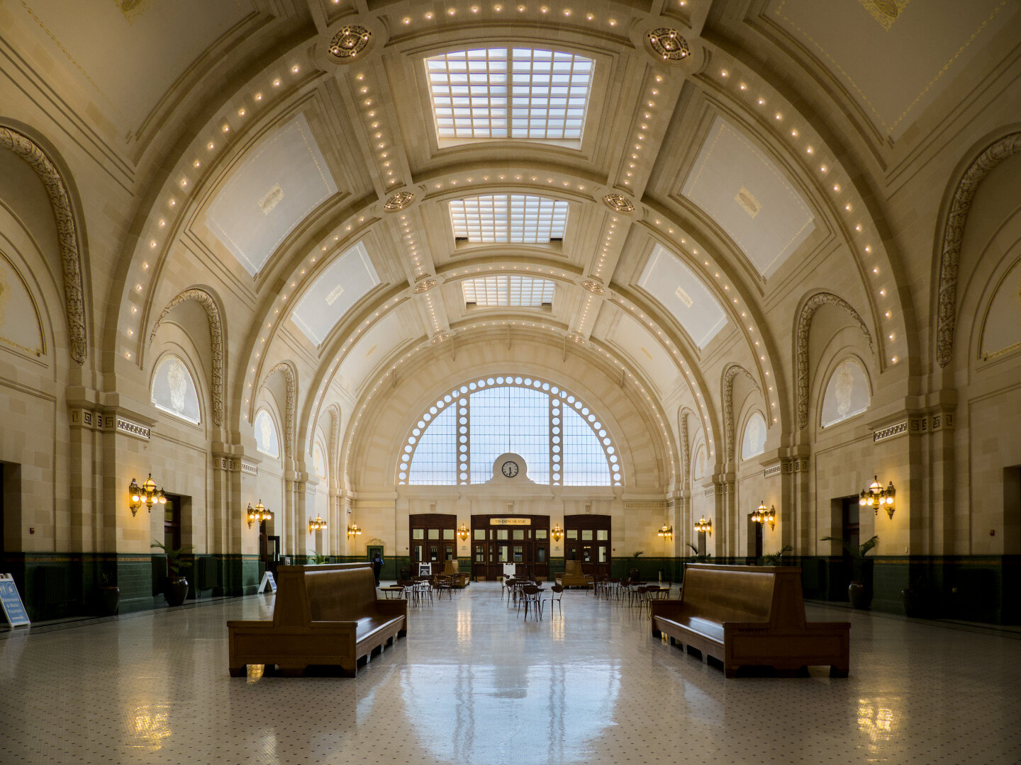 A grand semicircular Beaux Art vault illuminated by skylights. The vault dwarfs the wooden benches and tables inside. The white floor reflects the skylights and the semicircular window at the end.