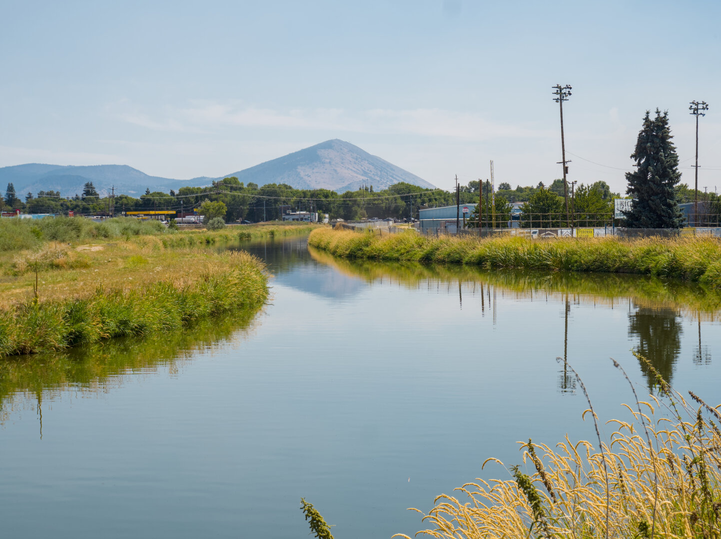 A wide and calm canal is flanked by mostly yellow grass, leading to a rounded mountains in the distance. The mountain, low-rise buildings nearby, and tall light poles are reflected in the canal.