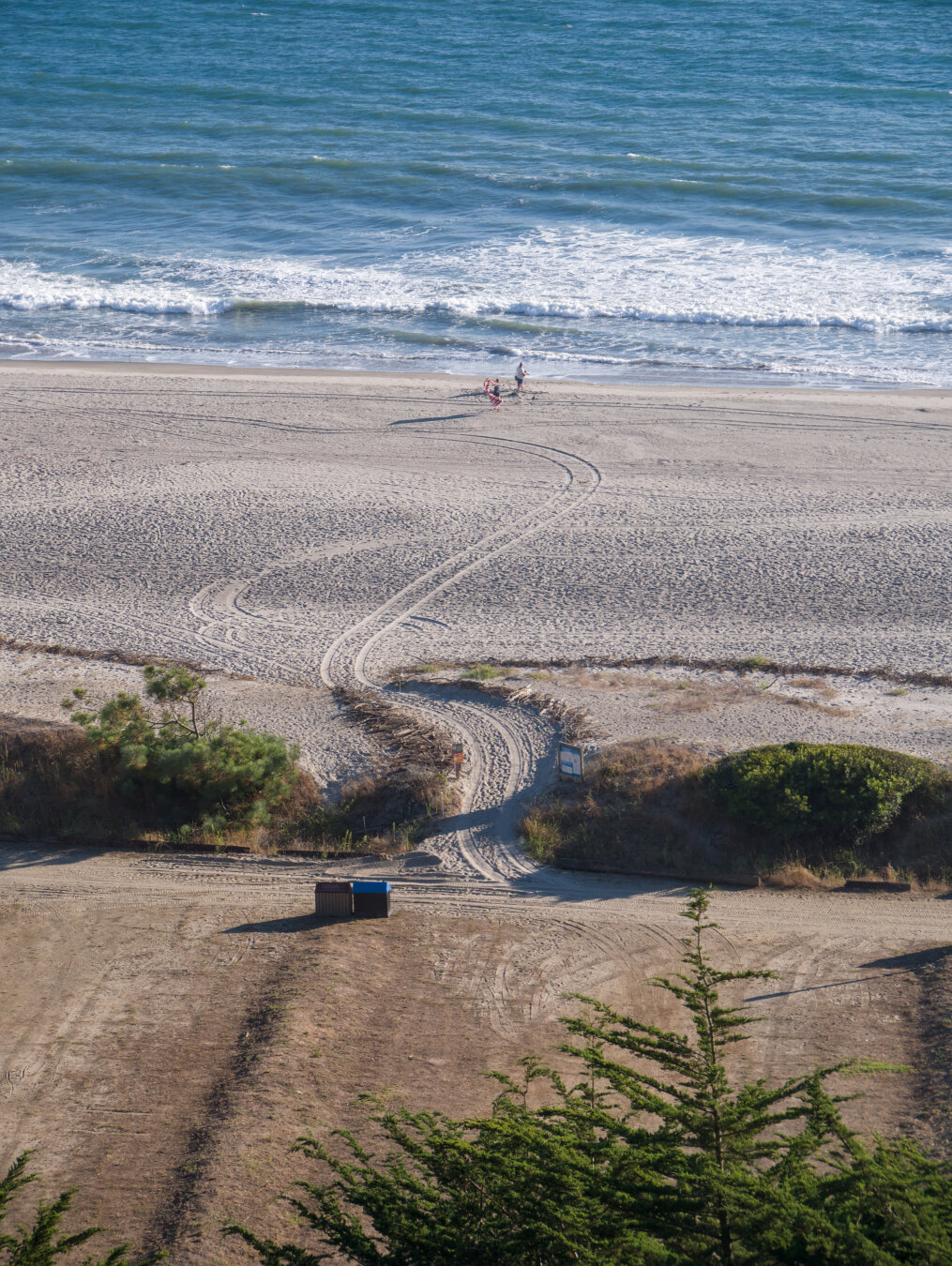 Looking down at a beach that has curvy tire treads leading to two people standing by the ocean