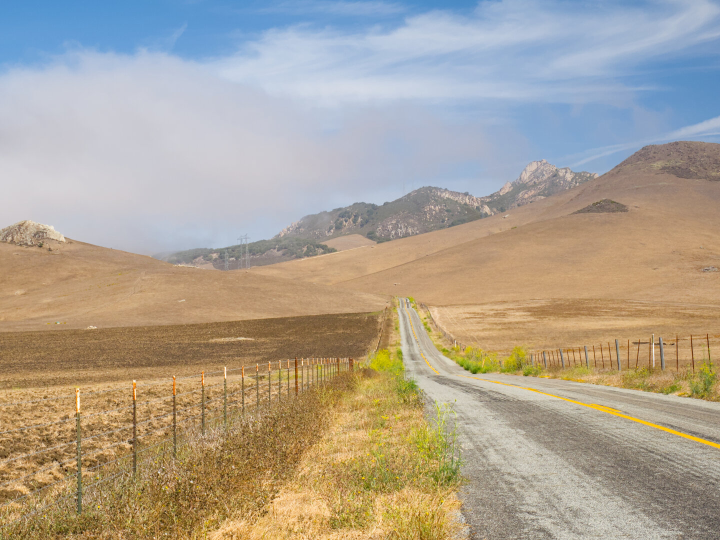 A small road with bad pavement leads into the scene towards rugged mountains with rocky tops but mostly covered by dry grass. If you look closely, you will see a cyclist in the distance, riding on the road.