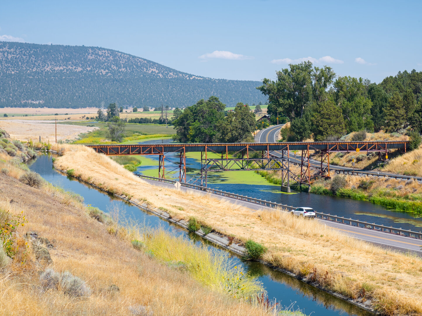 A narrow canal somewhat above a wider natural rider in an arid landscape; the river has some green algae and there's a small railroad bridge over the river. On the other side of the river is a small paved road between some trees. In the distance is a mountain covered in trees.