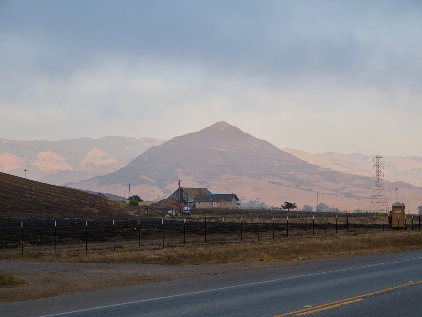A smaller conical mountain in front of a larger and wider mountain were illuminated by the golden afternoon sun. The mountains were largely covered in dry yellow grass and have green shrubs at higher altitudes and in the valleys. The mountains were just below dark clouds. At the foothills is farmlands and barns in the shade. There's a brown portapotty and a power line pylon on the right. There's a paved road in the foreground.