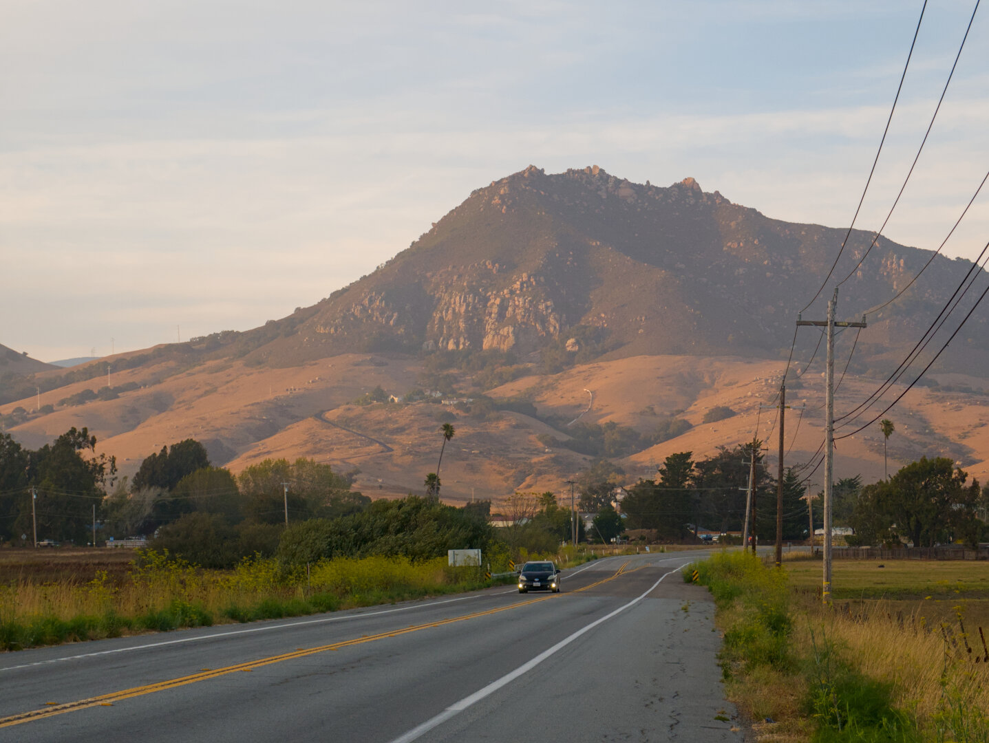 A 2 lane paved road leads to a great mountain basking in the golden setting sun. The mountain has gentler slopes covered in dry yellow grass and valleys with green shrubs at the bottom, and rugged rocks and steep cliffs at the top. There's a car traveling on the road towards me.