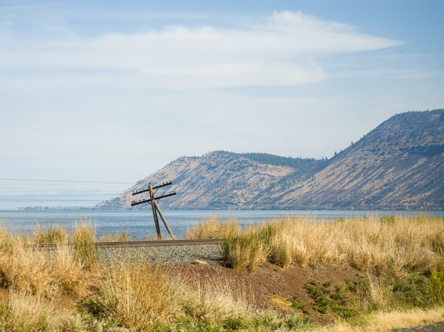 Two mountains by a vast lake, with a railroad track in the midst of dry grass and a wooden power line pole in the foreground