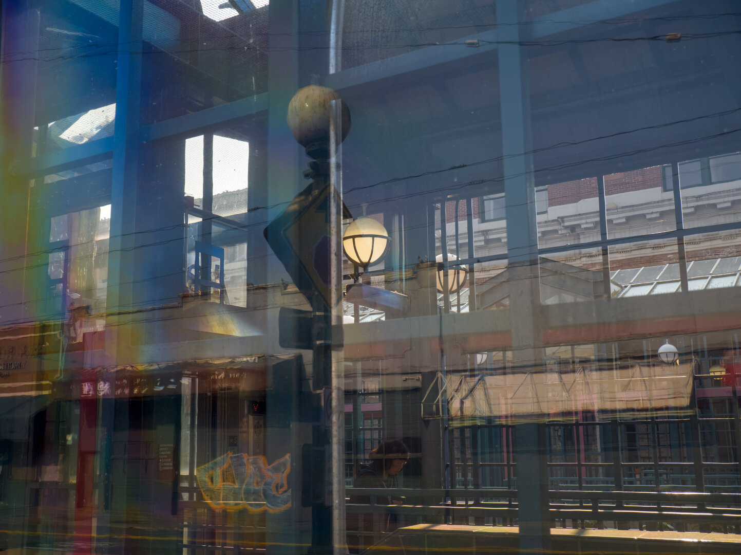 Reflections of the street scene by the glass wall are superimposed onto the interior of a modern train station. A person can be vaguely seen inside.