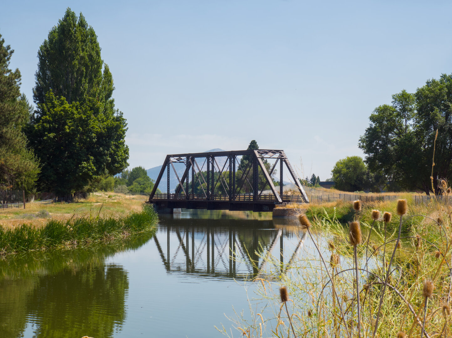 A small steel truss railroad bridge crosses a calm canal. The canal reflects the bridge and the taller trees around it.