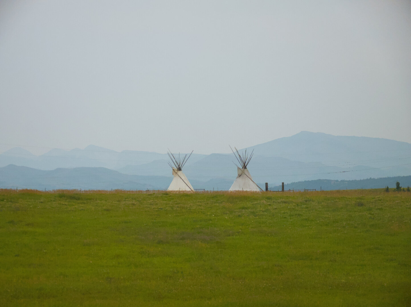 Two white teepee tends on vast green grassland, with mountains in the mist in the distance. It was cloudy.
