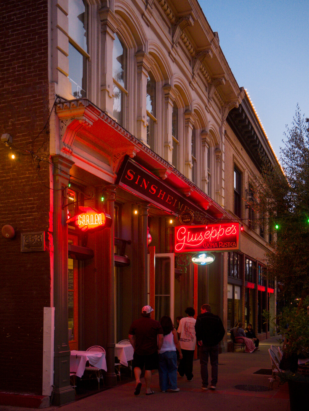 Looking on the side of a 2 story building with a brick wall facing an alley and a white Victorian style facade. There's an Italian restaurant on the 1st floor, with red neon signs. 4 people were walking by the restaurant.