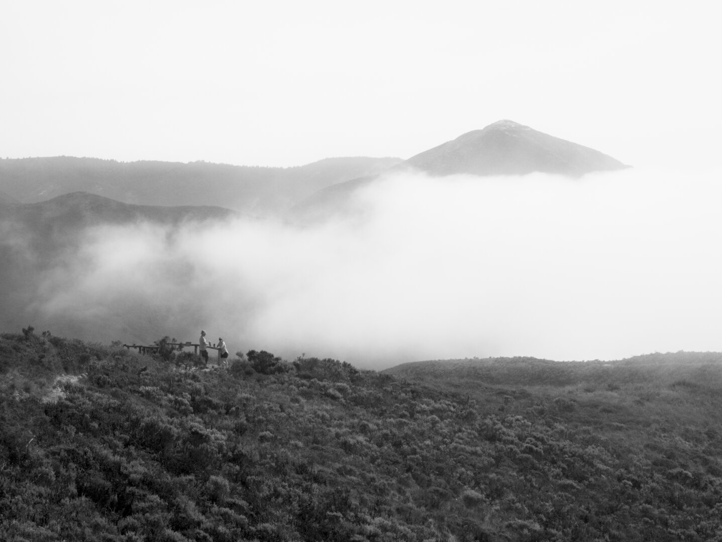 Clouds fill the valley between two mountains. On my side of the valley, there's a little scenic overlook with two people on it, dwarfed by the scene.
